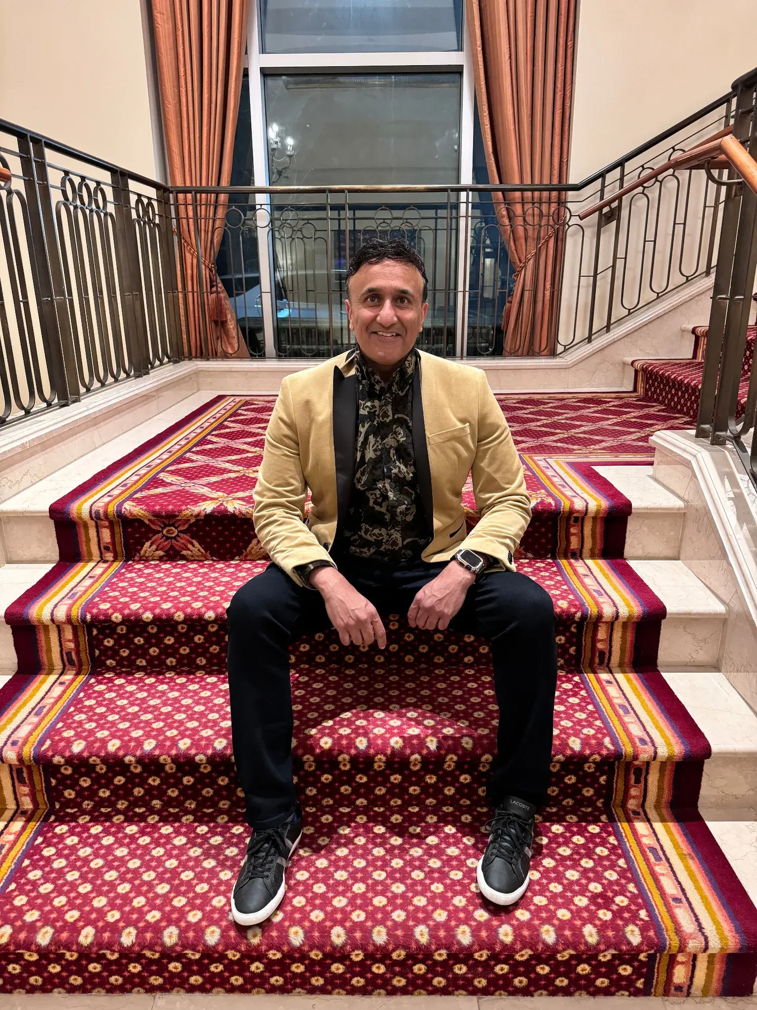 Neo smiling while seated on a red-carpeted staircase in a gold blazer